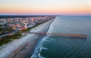 Aerial view of the Ocean Isle Beach Pier reaching into the Atlantic, with the beach and town along the shore under a pink and orange sky