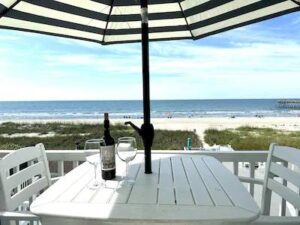 Outdoor dining table under a striped umbrella with a bottle of wine, with dune grass and the ocean beyond